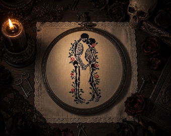 May include: A circular embroidery hoop containing a cross-stitch design of two skeletons holding hands and kissing, surrounded by red roses and green vines on a light beige fabric background, framed by a dark wooden hoop with a metal tension screw.