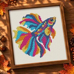 May include: A framed cross-stitch embroidery featuring a vibrant betta fish with flowing fins in shades of blue, pink, yellow, and orange. The fish is centred on a white Aida cloth background, enclosed within a simple, square wooden frame.