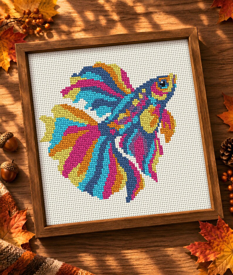 May include: A framed cross-stitch embroidery featuring a vibrant betta fish with flowing fins in shades of blue, pink, yellow, and orange. The fish is centred on a white Aida cloth background, enclosed within a simple, square wooden frame.