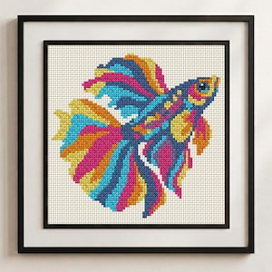 May include: A framed cross-stitch embroidery featuring a vibrant betta fish with flowing fins in shades of bright pink, electric blue, and sunny yellow against a white grid fabric background, enclosed within a simple black square frame.