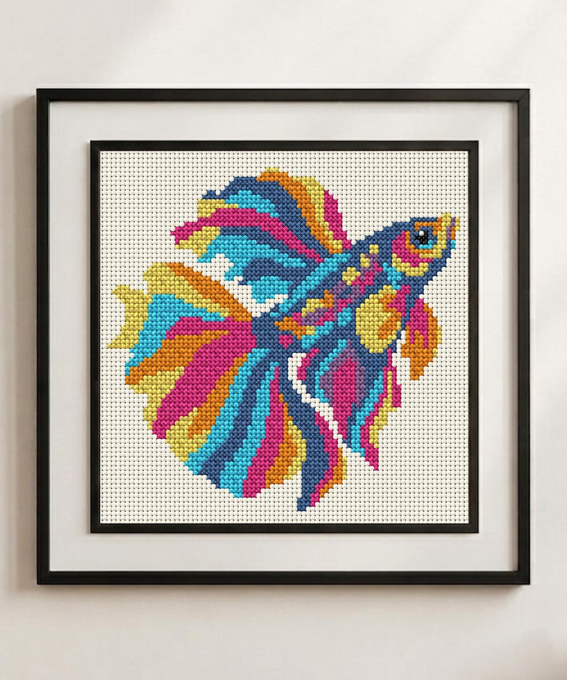 May include: A framed cross-stitch embroidery featuring a vibrant betta fish with flowing fins in shades of bright pink, electric blue, and sunny yellow against a white grid fabric background, enclosed within a simple black square frame.