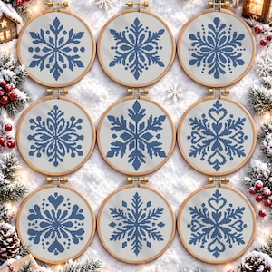 May include: Nine circular wooden embroidery hoops containing white fabric with blue cross-stitched snowflake patterns. Each hoop features a unique, symmetrical snowflake design centered on the fabric, arranged in a three-by-three grid.