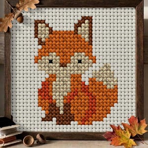 May include: A cross-stitch embroidery of a fox in shades of orange, cream, and dark brown, set against a white grid fabric. The piece is enclosed in a dark wooden frame, with an oak leaf and acorn accent attached to the top left corner.