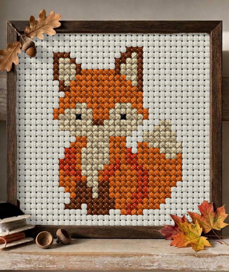 May include: A cross-stitch embroidery of a fox in shades of orange, cream, and dark brown, set against a white grid fabric. The piece is enclosed in a dark wooden frame, with an oak leaf and acorn accent attached to the top left corner.
