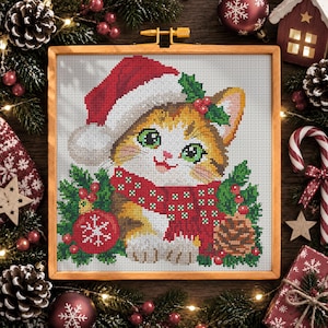 May include: A cross-stitch embroidery in a wooden frame depicting a kitten wearing a red and white Santa hat and a red patterned scarf. The kitten is surrounded by green holly leaves, red berries, a red Christmas ornament, and a pinecone.