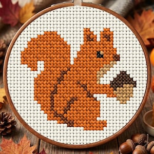 May include: A circular wooden embroidery hoop containing a cross-stitch pattern of a squirrel with orange and brown fur holding a brown acorn. The design is stitched onto a white fabric grid, featuring a small black eye and a tan chest area.