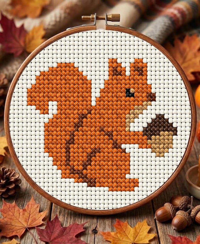 May include: A circular wooden embroidery hoop containing a cross-stitch pattern of a squirrel with orange and brown fur holding a brown acorn. The design is stitched onto a white fabric grid, featuring a small black eye and a tan chest area.
