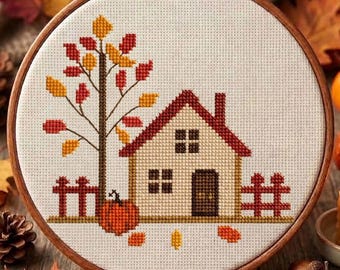 May include: A circular wooden embroidery hoop containing a cross-stitch design on white fabric. The pattern features a house with a red roof, a tree with red and yellow autumn leaves, a pumpkin, and a red fence, all rendered in pixelated style.