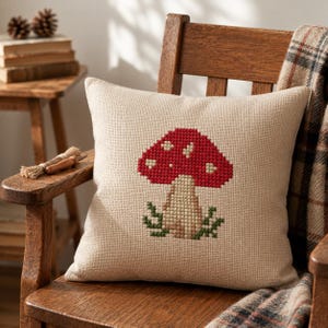 May include: A square throw pillow made of beige textured fabric featuring a centered cross-stitch design of a red mushroom with white spots and a light brown stem, surrounded by small green foliage at the base of the mushroom stalk.