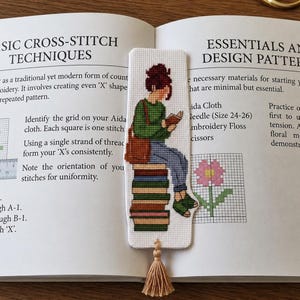 May include: A rectangular cross-stitch bookmark featuring a person with a brown bun hairstyle, wearing a green sweater, blue jeans, and green sandals, sitting on a stack of colorful books while reading. The bookmark has a beige tassel at the bottom.