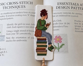 May include: A rectangular cross-stitch bookmark featuring a person with a brown bun hairstyle, wearing a green sweater, blue jeans, and green sandals, sitting on a stack of colorful books while reading. The bookmark has a beige tassel at the bottom.