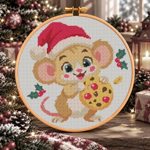 Op de afbeelding: Een kruissteekborduurwerk in een ronde houten ring met een beige muis die een rood-witte kerstmuts draagt. De muis houdt een geel koekje met rode en bruine stippen vast. Kleine groene hulstblaadjes met rode bessen omringen de muis.