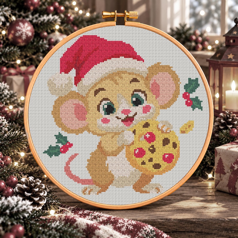 Op de afbeelding: Een kruissteekborduurwerk in een ronde houten ring met een beige muis die een rood-witte kerstmuts draagt. De muis houdt een geel koekje met rode en bruine stippen vast. Kleine groene hulstblaadjes met rode bessen omringen de muis.