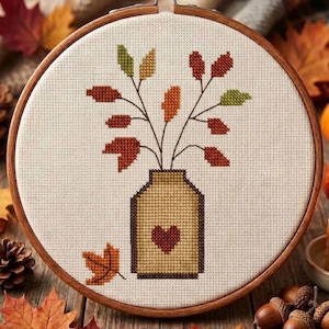 May include: A circular wooden embroidery hoop containing a cross-stitch pattern on off-white fabric. The design features a tan jar with a dark red heart, holding branches with autumn-coloured leaves in shades of red, orange, and green.
