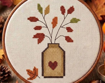 May include: A circular wooden embroidery hoop containing a cross-stitch pattern on off-white fabric. The design features a tan jar with a dark red heart, holding branches with autumn-colored leaves in shades of red, orange, and green.