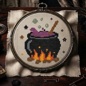 May include: A cross-stitch embroidery piece in a round, ornate bronze-coloured frame. The design features a black cauldron filled with purple liquid, a wooden stirring stick, and orange flames at the base, set against a light beige fabric background.