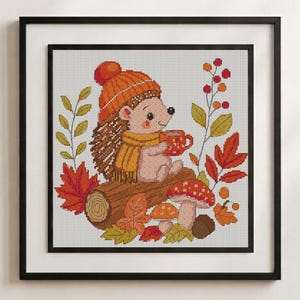 May include: A framed cross-stitch embroidery featuring a hedgehog wearing an orange knit hat and yellow scarf, holding a red mug. The hedgehog sits on a brown log surrounded by red and yellow autumn leaves, two spotted mushrooms, and berries.