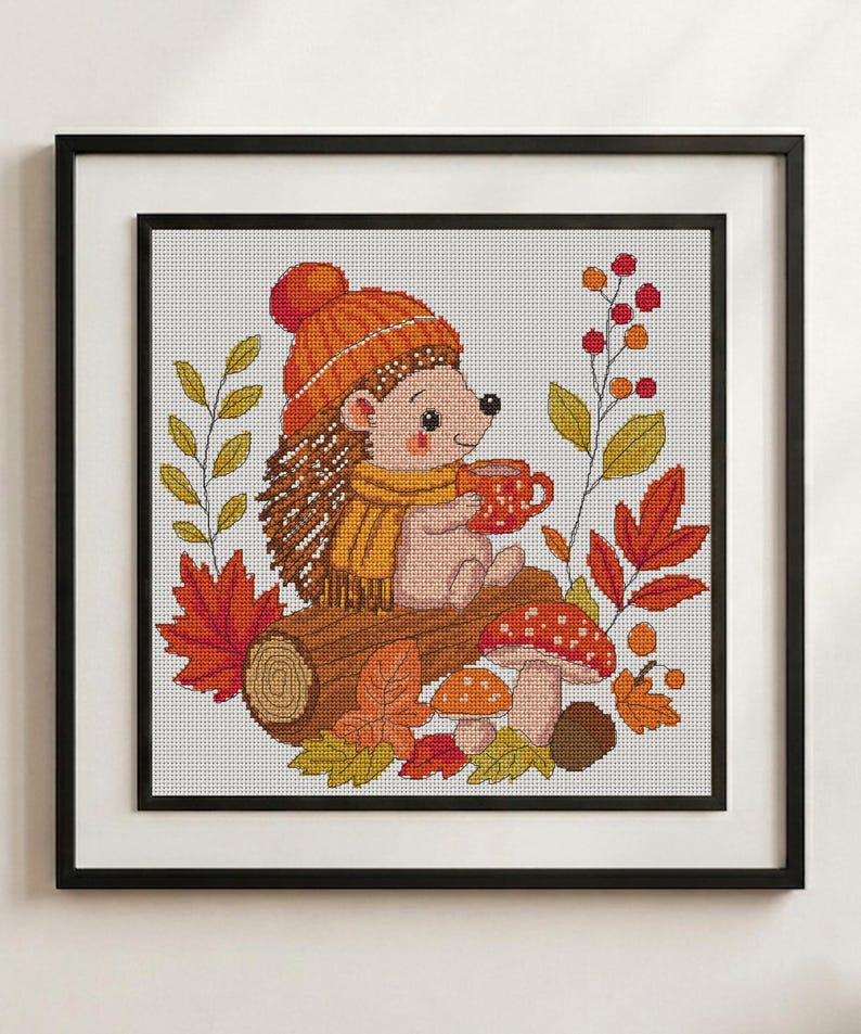 May include: A framed cross-stitch embroidery featuring a hedgehog wearing an orange knit hat and yellow scarf, holding a red mug. The hedgehog sits on a brown log surrounded by red and yellow autumn leaves, two spotted mushrooms, and berries.