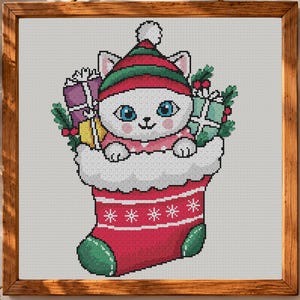 May include: A cross-stitch pattern in a wooden frame featuring a white cat with blue eyes wearing a red and green striped hat. The cat is inside a red Christmas stocking with white trim, surrounded by wrapped gifts and green holly leaves.