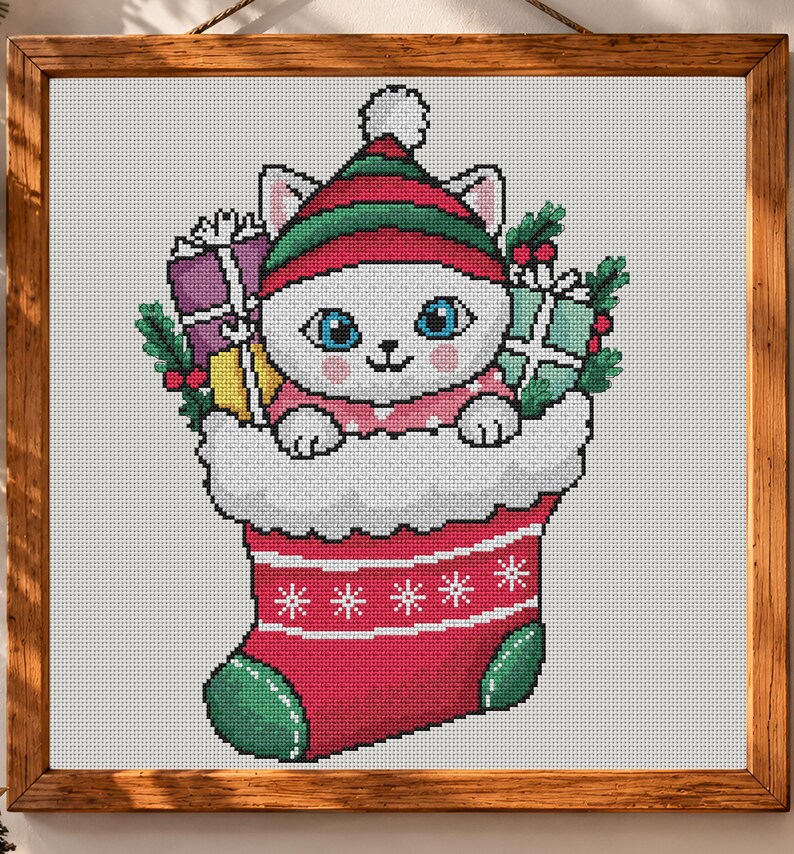 May include: A cross-stitch pattern in a wooden frame featuring a white cat with blue eyes wearing a red and green striped hat. The cat is inside a red Christmas stocking with white trim, surrounded by wrapped gifts and green holly leaves.