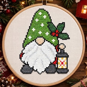 May include: A cross-stitch pattern in a wooden embroidery hoop featuring a gnome with a large white beard, wearing a green hat with white cross-shaped accents, decorated with holly leaves and red berries, holding a small lantern with a red center.