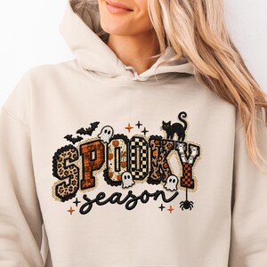 May include: A beige hooded sweatshirt featuring a graphic print with the text "SPOOKY season" in black and orange. The letters are filled with various patterns including leopard print, plaid, and pumpkins, surrounded by ghosts, bats, and a cat.