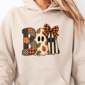 May include: A beige hooded sweatshirt featuring a graphic print of the word BOO. The letter B is patterned with black and orange plaid and leopard print. Two ghost figures follow, one covered in autumn leaves and one with black stripes and an orange plaid bow.