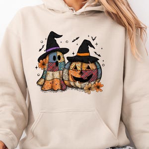 May include: A beige hooded sweatshirt featuring a graphic print of a patchwork ghost and a patchwork jack-o'-lantern, both wearing black witch hats. The design includes small orange flowers, bats, and stars, rendered in a colorful, quilted pattern.