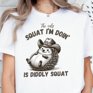 May include: A white t-shirt featuring a graphic of a hedgehog wearing a cowboy hat. The text above the hedgehog reads The only SQUAT I'M DOIN' and the text below reads IS DIDDLY SQUAT. The design is rendered in shades of brown and black ink.
