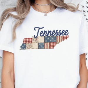 Op de afbeelding: Een wit t-shirt met korte mouwen met het woord Tennessee in donkerblauw schrift boven een afbeelding van de staat Tennessee, gevuld met een patchworkpatroon van rood-wit gingham, blauw met witte sterren en bloemenprints.