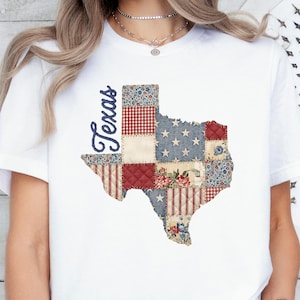 May include: A white short-sleeved t-shirt featuring a graphic of the state of Texas. The state shape is filled with a patchwork design of various patterns including red and white gingham, blue with white stars, and floral prints. The word Texas is printed in blue script to the left.