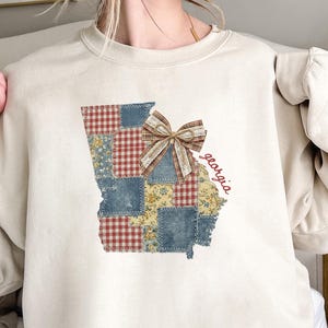 Puede incluir: Una sudadera de cuello redondo color crema con un gráfico del estado de Georgia. La forma del estado está rellena con un patrón de retazos de cuadros vichy rojos y blancos, mezclilla azul y tela floral amarilla, acentuada por un lazo de cinta a cuadros.