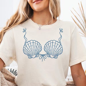 Può includere: Una t-shirt a maniche corte color crema con una grafica a linee blu scuro raffigurante due conchiglie disposte come un top bikini, completa di sottili dettagli a filo che collegano le conchiglie e si estendono verso l'alto come spalline.