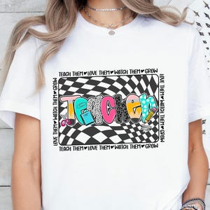 May include: A white t-shirt featuring a central graphic with a black and white checkered background. The word TEACHER is written in colourful, patterned letters. The text TEACH THEM LOVE THEM WATCH THEM GROW surrounds the rectangular design.