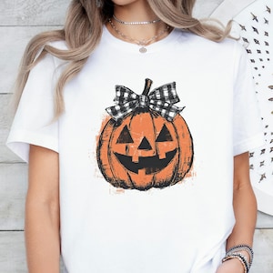 May include: A white short-sleeve t-shirt featuring a central graphic of an orange jack-o'-lantern pumpkin with a black and white buffalo plaid bow on top. The pumpkin has a distressed, textured appearance with black triangular eyes and a wide grin.