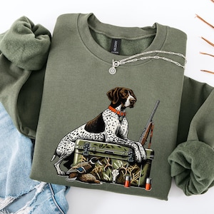 Op de afbeelding: Een olijfgroen sweatshirt met ronde hals en een afbeelding van een wit met bruin gevlekte jachthond die op een camouflage koelbox rust. Het ontwerp bevat een jachtgeweer, een eend, een hertenkop en twee jachtpatronen in detail.