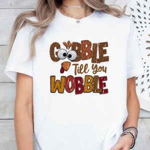 May include: A white short-sleeved t-shirt featuring a graphic print with the text GOBBLE Till you WOBBLE. The letters have a textured brown and orange pattern, and the letter O in GOBBLE is replaced by a cartoon turkey face with large eyes.