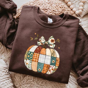 Può includere: Una felpa girocollo marrone scuro con una grafica centrale raffigurante una zucca composta da vari motivi patchwork nei toni dell'arancione, del verde e del bianco. La zucca è sormontata da un fiocco decorativo con stampa floreale e fogliame.
