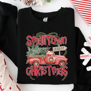 May include: A black crewneck sweatshirt featuring a graphic of a red pickup truck carrying a Christmas tree. The text reads Small Town Christmas and 77077 on a wooden sign. The design includes a wreath on the truck door and green ribbon accents.