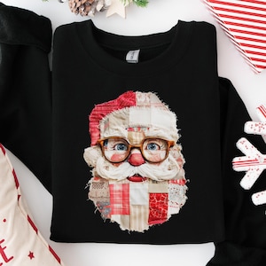 May include: A black long-sleeve sweatshirt featuring a central graphic of a Santa Claus face constructed from various red, white, and beige fabric patches. The Santa figure wears brown-rimmed glasses and has a prominent red nose and mustache.