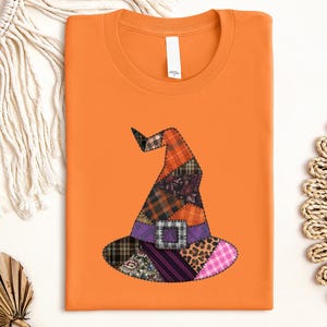 Puede incluir: Una camiseta de color naranja brillante con un gráfico central de un sombrero de bruja hecho de varios patrones de tela, incluyendo cuadros, estampado de leopardo, flores y vichy, con una banda morada y una hebilla a cuadros grises.
