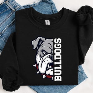 May include: A black crewneck sweatshirt featuring a graphic of a grey bulldog face wearing a red spiked collar, positioned next to the white text "BULLDOGS" in a bold, block font. The sweatshirt is layered over a pair of blue denim jeans.
