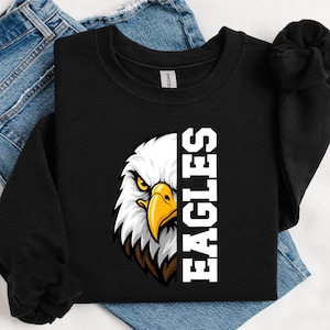 Può includere: Una felpa nera a maniche lunghe con la grafica della testa di un'aquila sul lato sinistro e la parola EAGLES stampata in grandi lettere bianche verticali sul lato destro. L'aquila ha piume bianche, un becco giallo e occhi scuri.