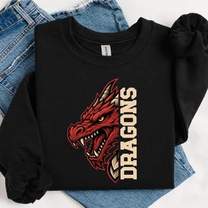 May include: A black long-sleeve sweatshirt featuring a graphic of a red dragon head with sharp teeth and yellow eyes, positioned next to the vertical text DRAGONS in a bold, light-coloured font. The garment has a crew neckline and ribbed cuffs.