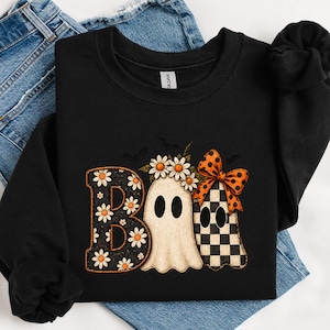 Peut inclure: Un sweat-shirt noir à manches longues avec une impression graphique du mot BOO. La lettre B a un fond noir avec des marguerites blanches, le O est un fantôme portant une couronne de marguerites, et le second O est un fantôme à carreaux avec un nœud orange à pois.