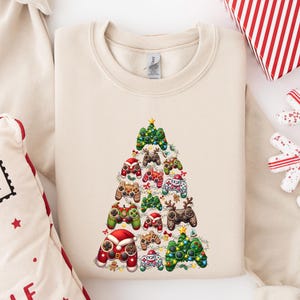May include: A beige crewneck sweatshirt featuring a graphic print of a Christmas tree constructed from various video game controllers. The controllers are decorated with holiday elements like Santa hats, reindeer antlers, and colourful string lights.