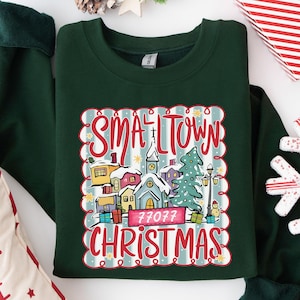 May include: A dark green crewneck sweatshirt featuring a graphic print of a snowy village scene with a church, houses, a Christmas tree, and gifts. The text reads "SMALL TOWN CHRISTMAS" in red script with the number "77077" on a pink rectangular banner.