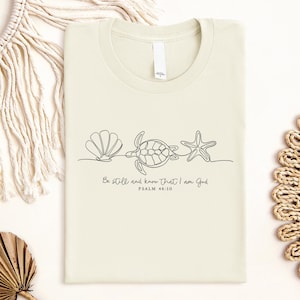 Könnte beinhalten: Ein cremefarbenes Kurzarm-T-Shirt mit einer schwarzen Strichzeichnung, die eine Muschel, eine Meeresschildkröte und einen Seestern zeigt. Unter der Illustration steht der Text: Be still and know that I am God, PSALM 46:10. Mit Rundhalsausschnitt.