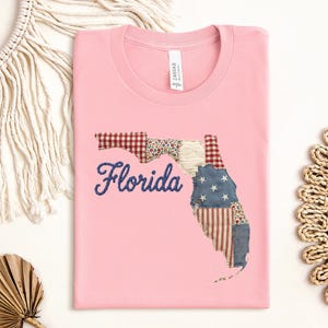 May include: A light pink t-shirt featuring a central graphic of the state of Florida. The state shape is filled with a patchwork design of red and white gingham, blue with white stars, and red and white stripes. The word Florida is embroidered in blue.