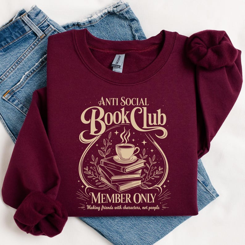 May include: A maroon crewneck sweatshirt featuring a cream-coloured graphic with the text "ANTI SOCIAL BOOK CLUB MEMBER ONLY" and "Making friends with characters, not people." The design includes a steaming cup of coffee resting on a stack of books.